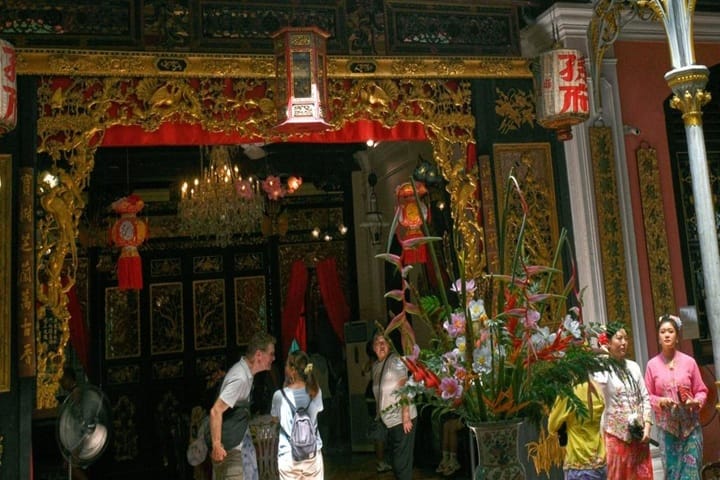 Interior of an ornate Asian temple with gold carvings, red curtains, and a large floral arrangement; several visitors walk through a decorated doorway.