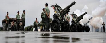 Soldiers in green uniforms stand beside large artillery cannons firing during a military parade on a wet surface.