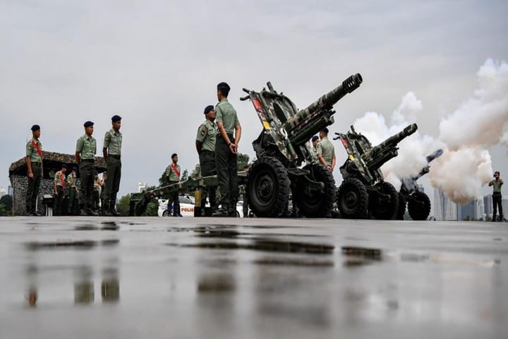 Soldiers in green uniforms stand beside large artillery cannons firing during a military parade on a wet surface.