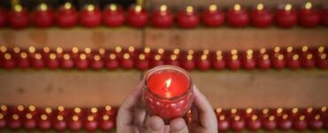 Hands cradle a lit red candle in a glass holder, with rows of red candles lining wooden steps in the background.