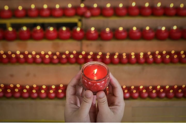 Hands cradle a lit red candle in a glass holder, with rows of red candles lining wooden steps in the background.