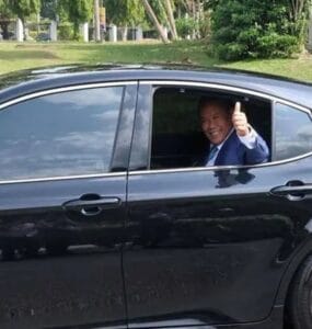 Smiling man in a blue suit giving a thumbs-up from the driver's window of a black sedan on a sunny day.