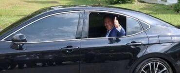Smiling man in a blue suit giving a thumbs-up from the driver's window of a black sedan on a sunny day.