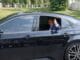 Smiling man in a blue suit giving a thumbs-up from the driver's window of a black sedan on a sunny day.