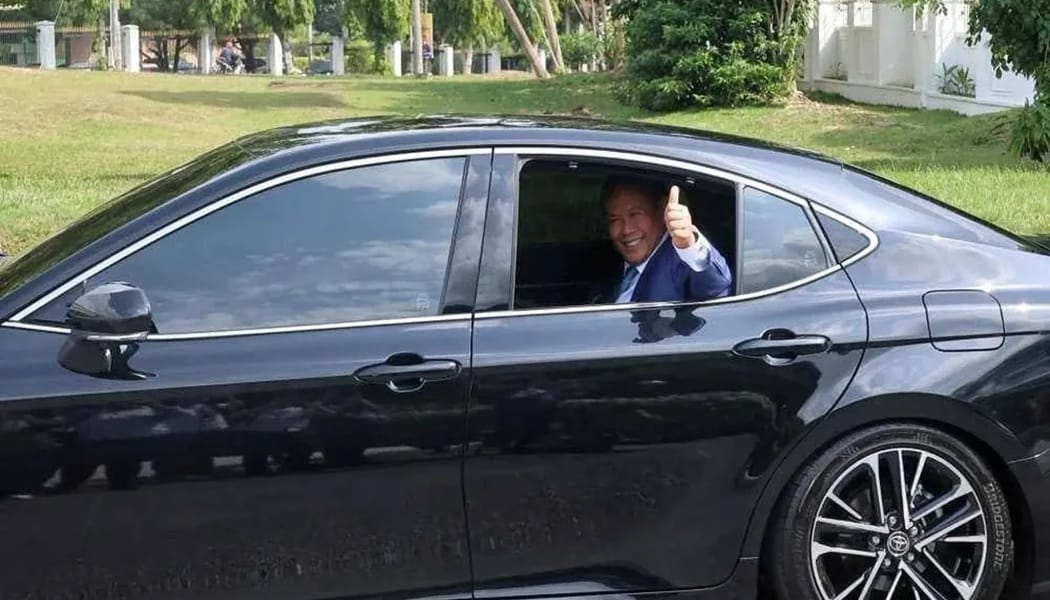 Smiling man in a blue suit giving a thumbs-up from the driver's window of a black sedan on a sunny day.