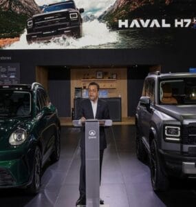 Man in suit at a podium presenting between two Haval SUVs in a showroom, with a large vehicle banner above.