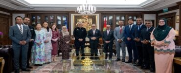 Group of professionals in formal attire posing for a photo in a wood-paneled meeting room with a chandelier and decorative rug.