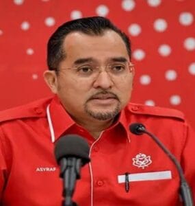 Man in a red uniform speaking at a press conference, with two microphones in front and a red dotted backdrop behind him.