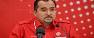 Man in a red uniform speaking at a press conference, with two microphones in front and a red dotted backdrop behind him.