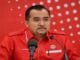 Man in a red uniform speaking at a press conference, with two microphones in front and a red dotted backdrop behind him.