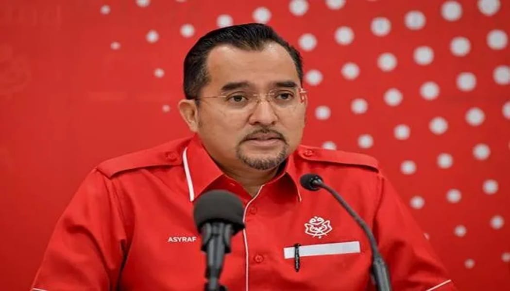 Man in a red uniform speaking at a press conference, with two microphones in front and a red dotted backdrop behind him.