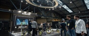 NAVEE exhibition booth at a trade show displaying electric bikes with bright yellow rims and a large screen backdrop.