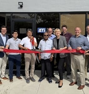 Group of people standing in a line outside a storefront, holding a red ribbon for a ribbon-cutting ceremony.