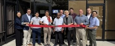 Group of people standing in a line outside a storefront, holding a red ribbon for a ribbon-cutting ceremony.
