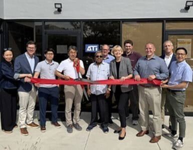 Group of people standing in a line outside a storefront, holding a red ribbon for a ribbon-cutting ceremony.