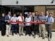 Group of people standing in a line outside a storefront, holding a red ribbon for a ribbon-cutting ceremony.