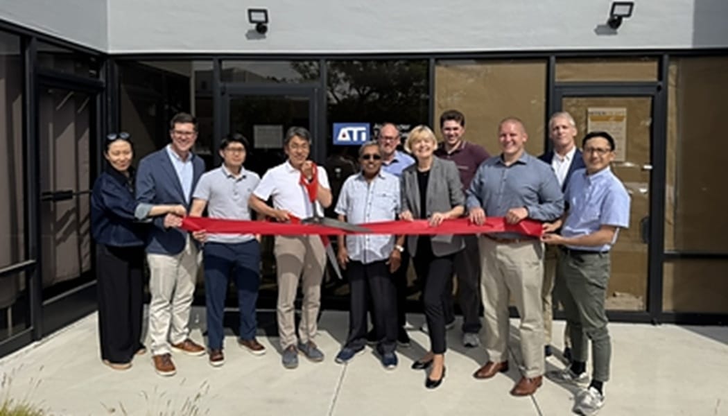 Group of people standing in a line outside a storefront, holding a red ribbon for a ribbon-cutting ceremony.