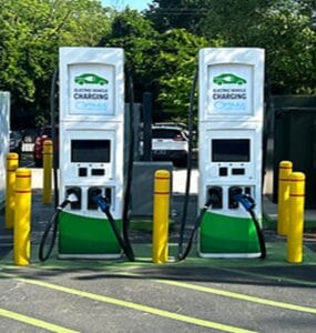 Two white-green electric vehicle charging stations in a parking lot with charging cables connected, flanked by yellow bollards.