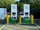 Two white-green electric vehicle charging stations in a parking lot with charging cables connected, flanked by yellow bollards.