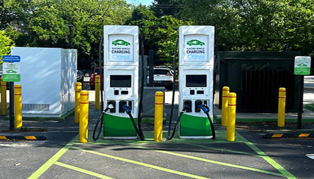 Two white-green electric vehicle charging stations in a parking lot with charging cables connected, flanked by yellow bollards.