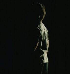 A person standing in a dark space, illuminated from the side, with their back turned to the viewer.