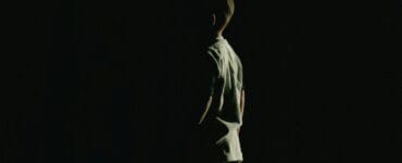 A person standing in a dark space, illuminated from the side, with their back turned to the viewer.