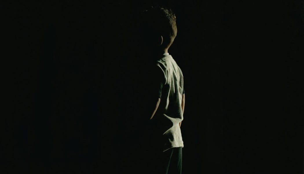 A person standing in a dark space, illuminated from the side, with their back turned to the viewer.